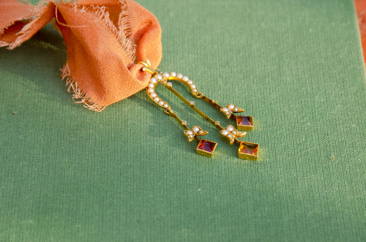 Gold pendant necklace with seed pearls and orange citrine drops on a textured green surface.