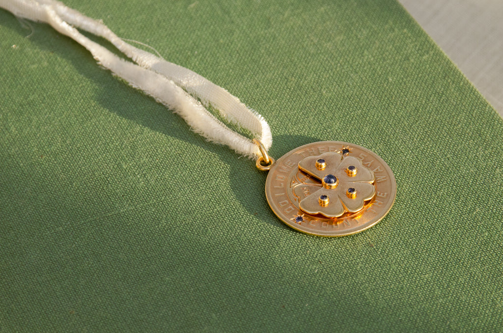A round gold pendant with a floral design and blue stone accents, resting on a textured green surface.