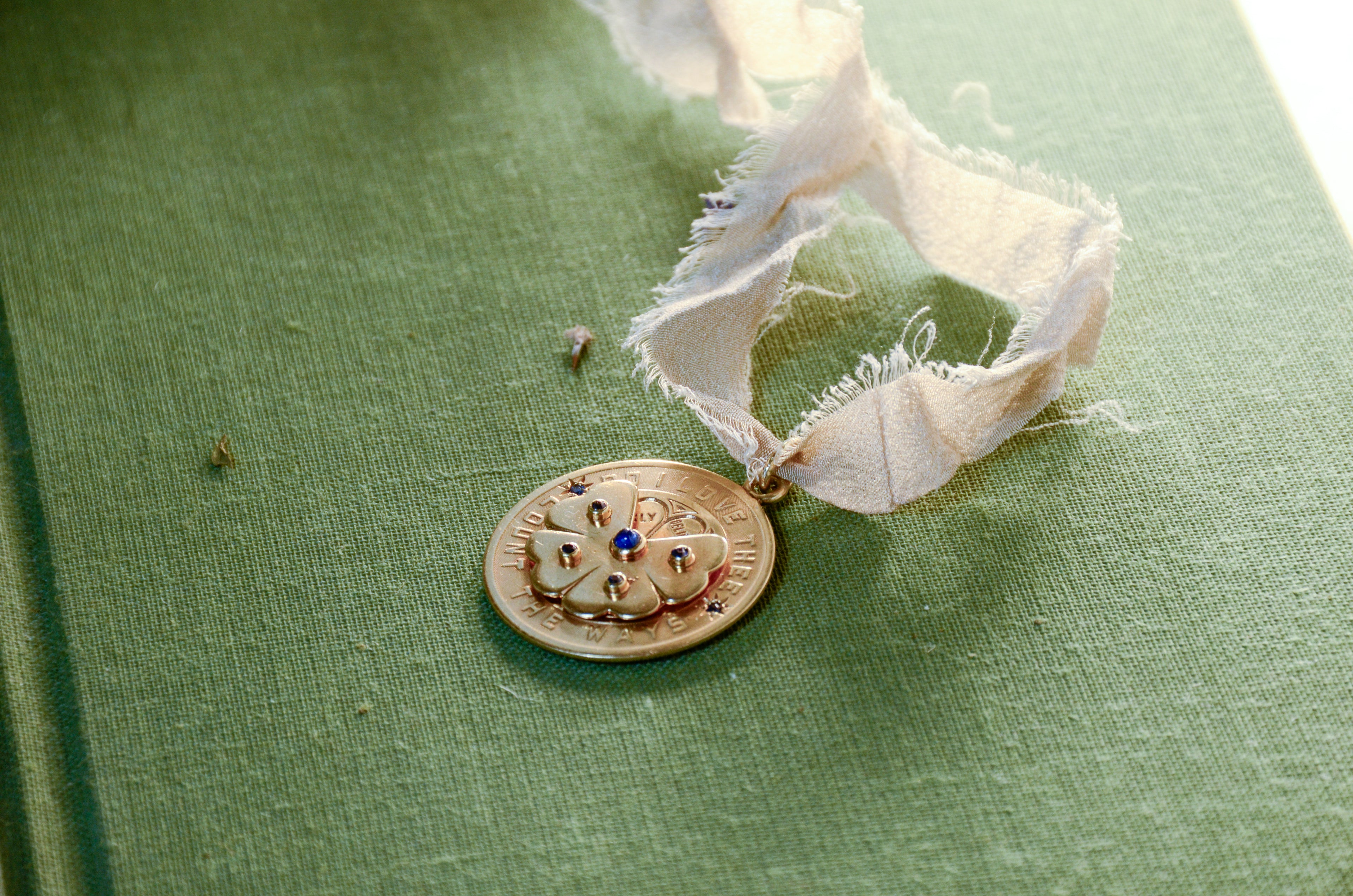 A round gold pendant with a floral design and blue stone accents, resting on a textured green surface.