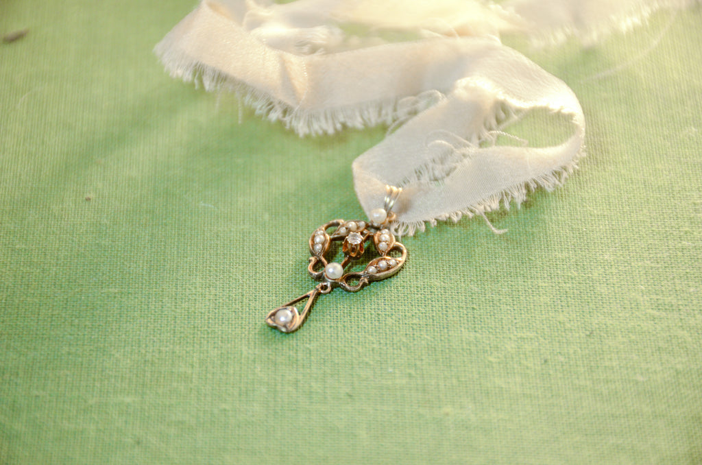 A gold lavalier pendant with seed pearls and a center diamond resting on a green textured surface.