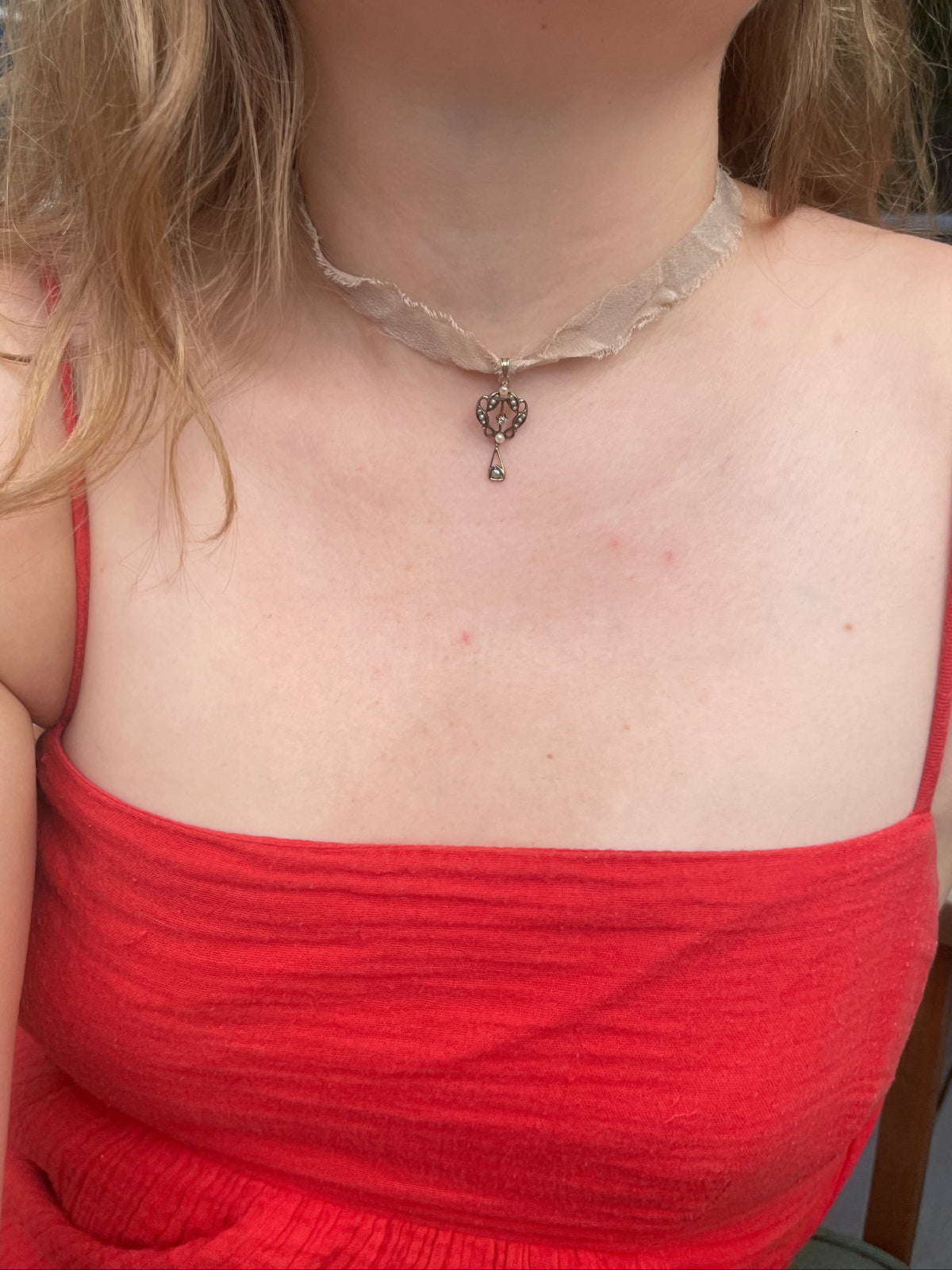 A close-up of a person wearing a delicate beige silk choker necklace with a small gold lavalier pendant covered in seed pearls and one center, diamond paired with a red ribbed camisole top.