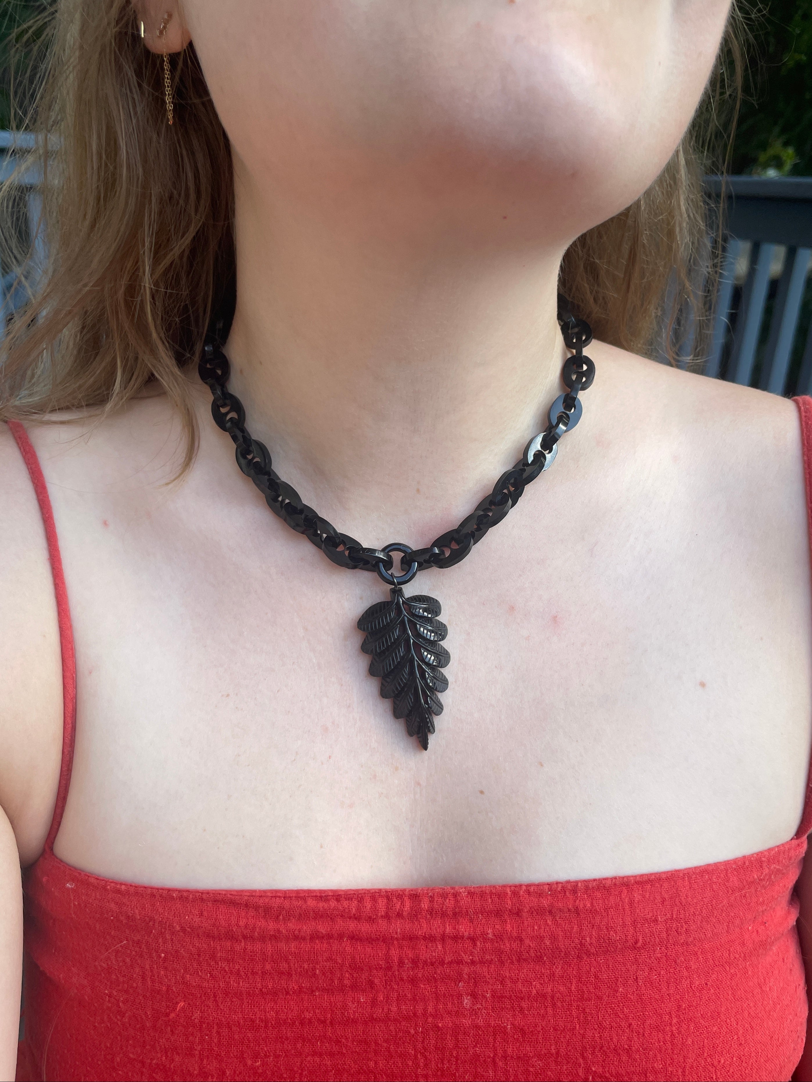 A close-up of a person wearing a dark charcoal gray beaded chain necklace with a matching leaf-shaped pendant, paired with a red spaghetti-strap top.