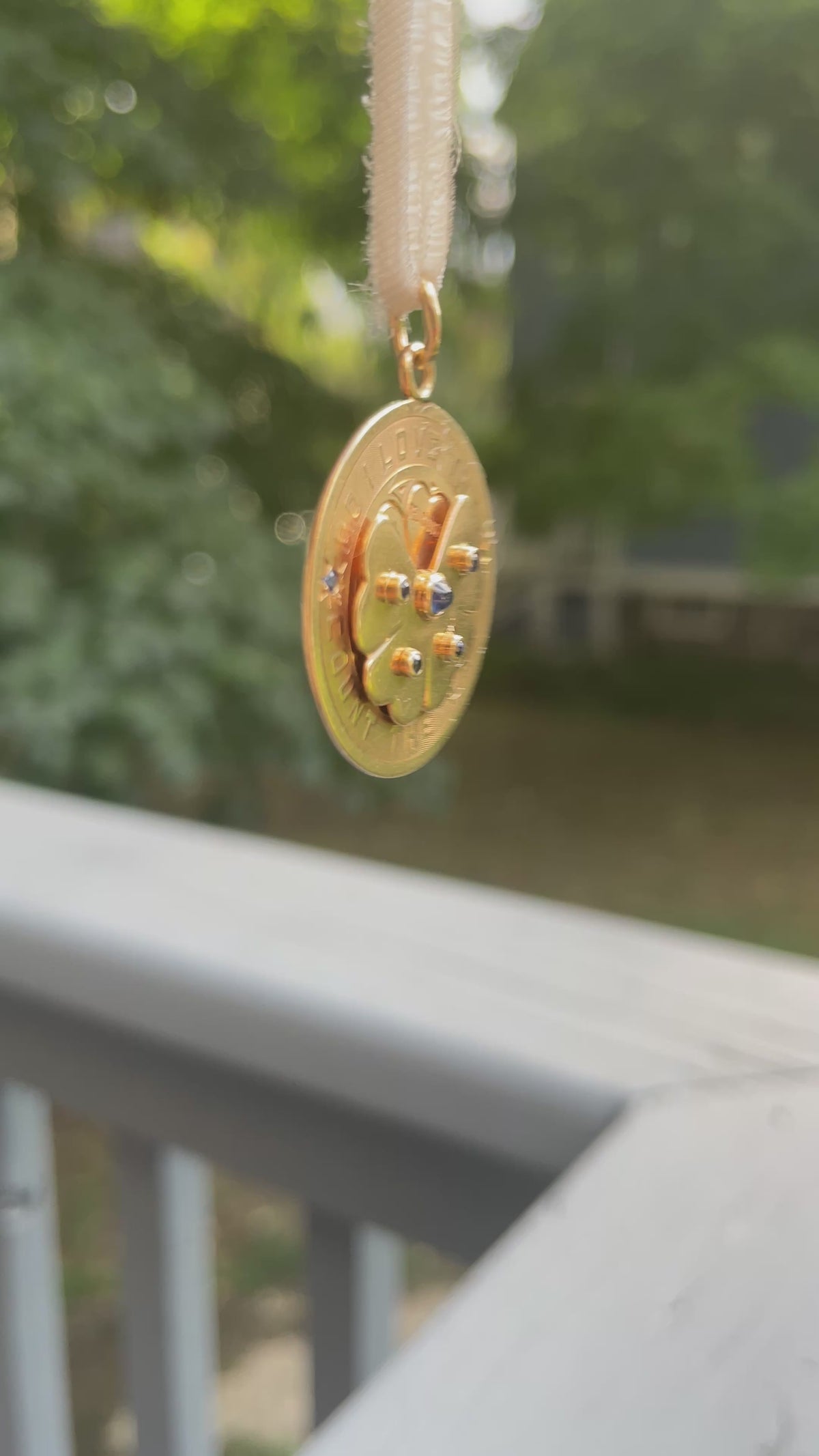 A round gold pendant with a floral design and blue stone accents, swaying on a white ribbon.