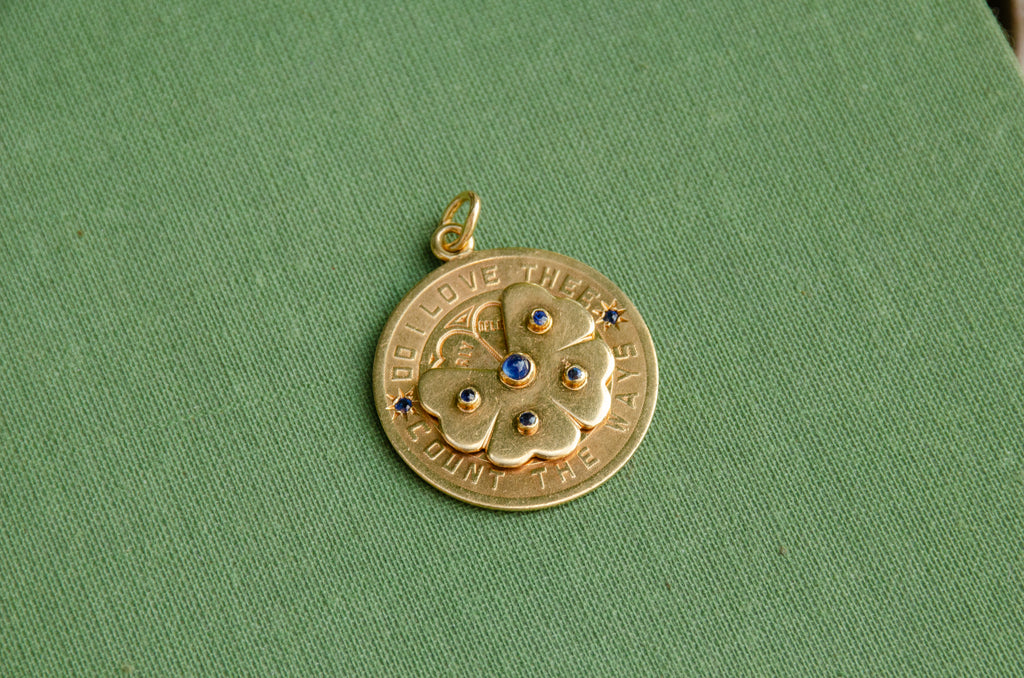 A round gold pendant with a floral design and blue stone accents, resting on a textured green surface.