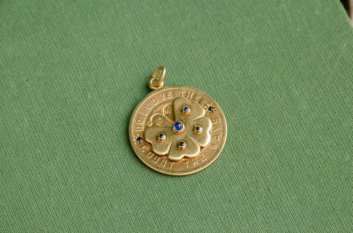 A round gold pendant with a floral design and blue stone accents, resting on a textured green surface.