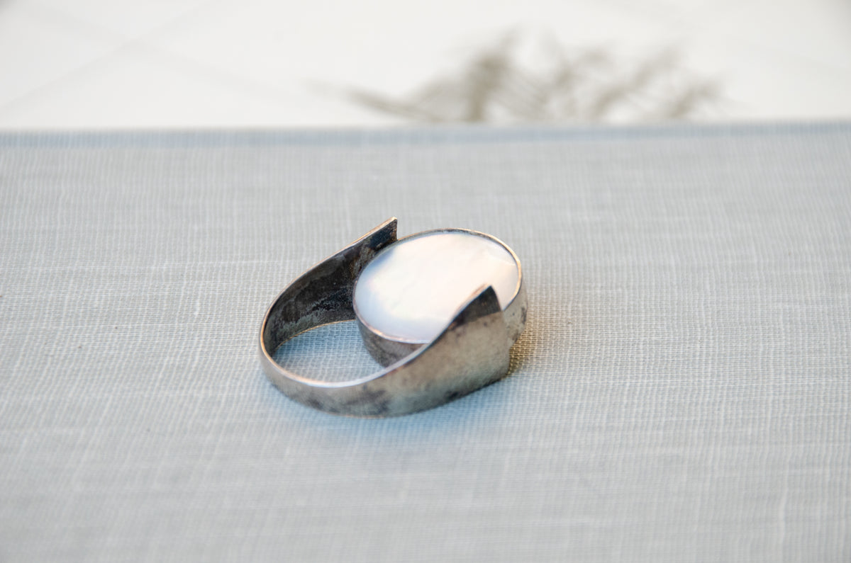 A silver ring with a large, smooth white oval mother of pearl stone, resting on a textured light gray fabric surface.