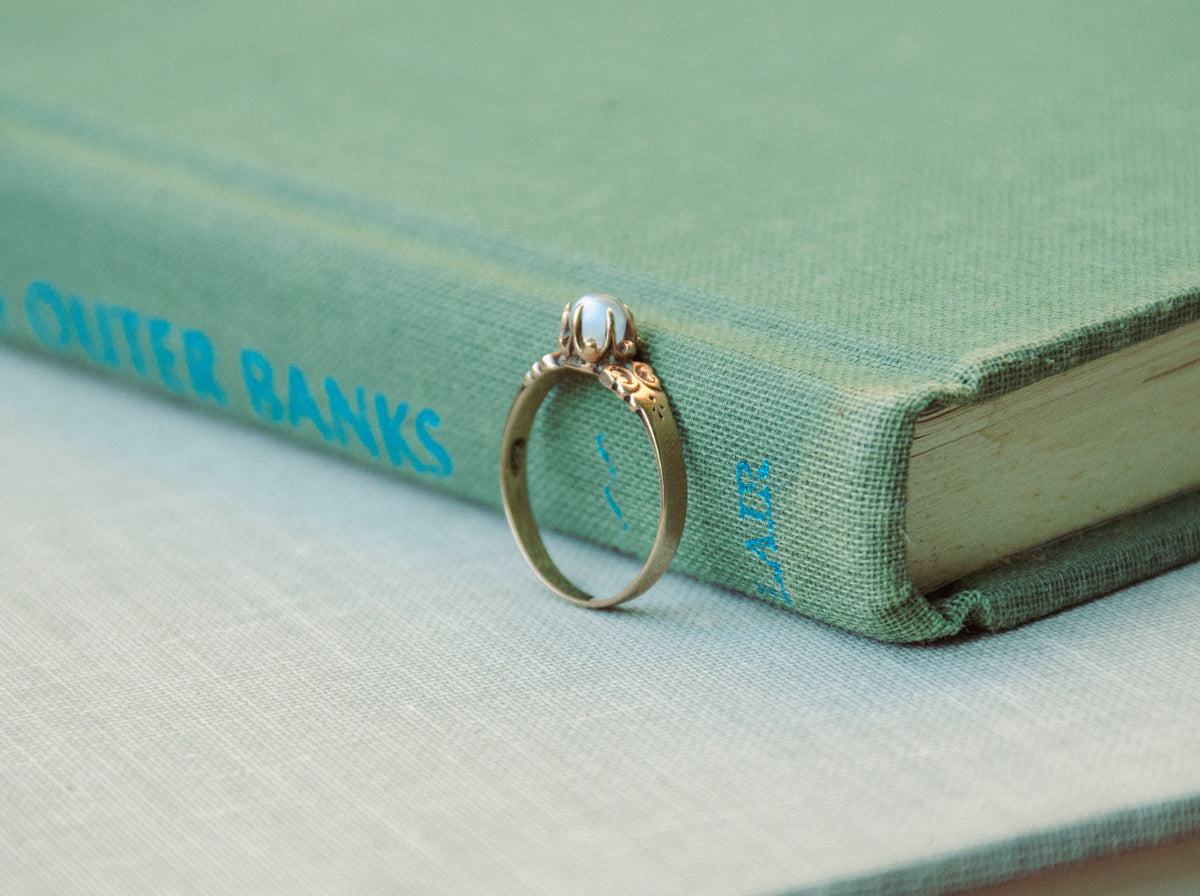 A rose gold ring with an ornate filigree design and a round light blue moonstone, resting on a textured sage green surface.
