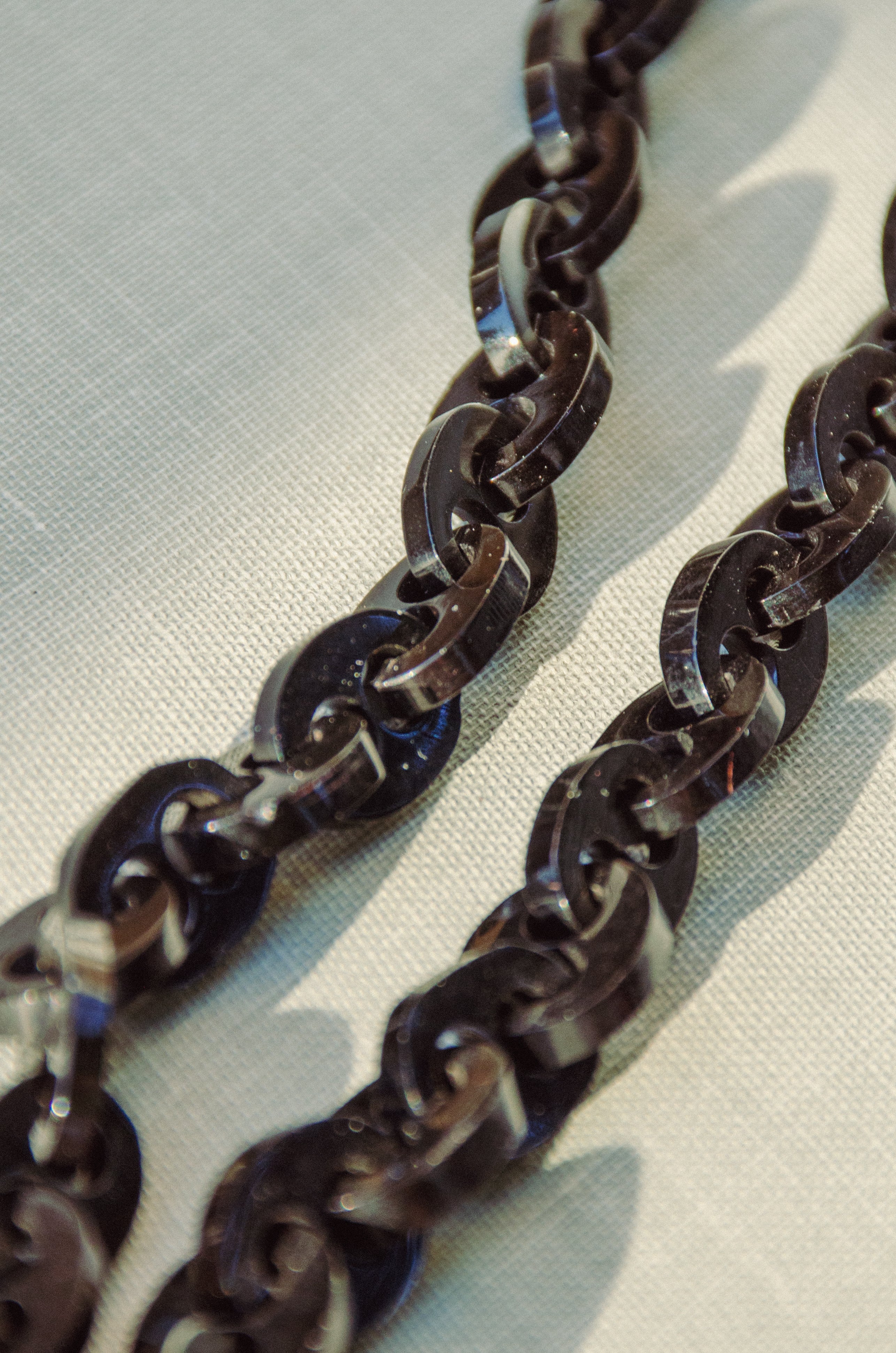 A close-up of a dark brown glossy chain necklace with interlocking links, resting on a light beige textured fabric background.
