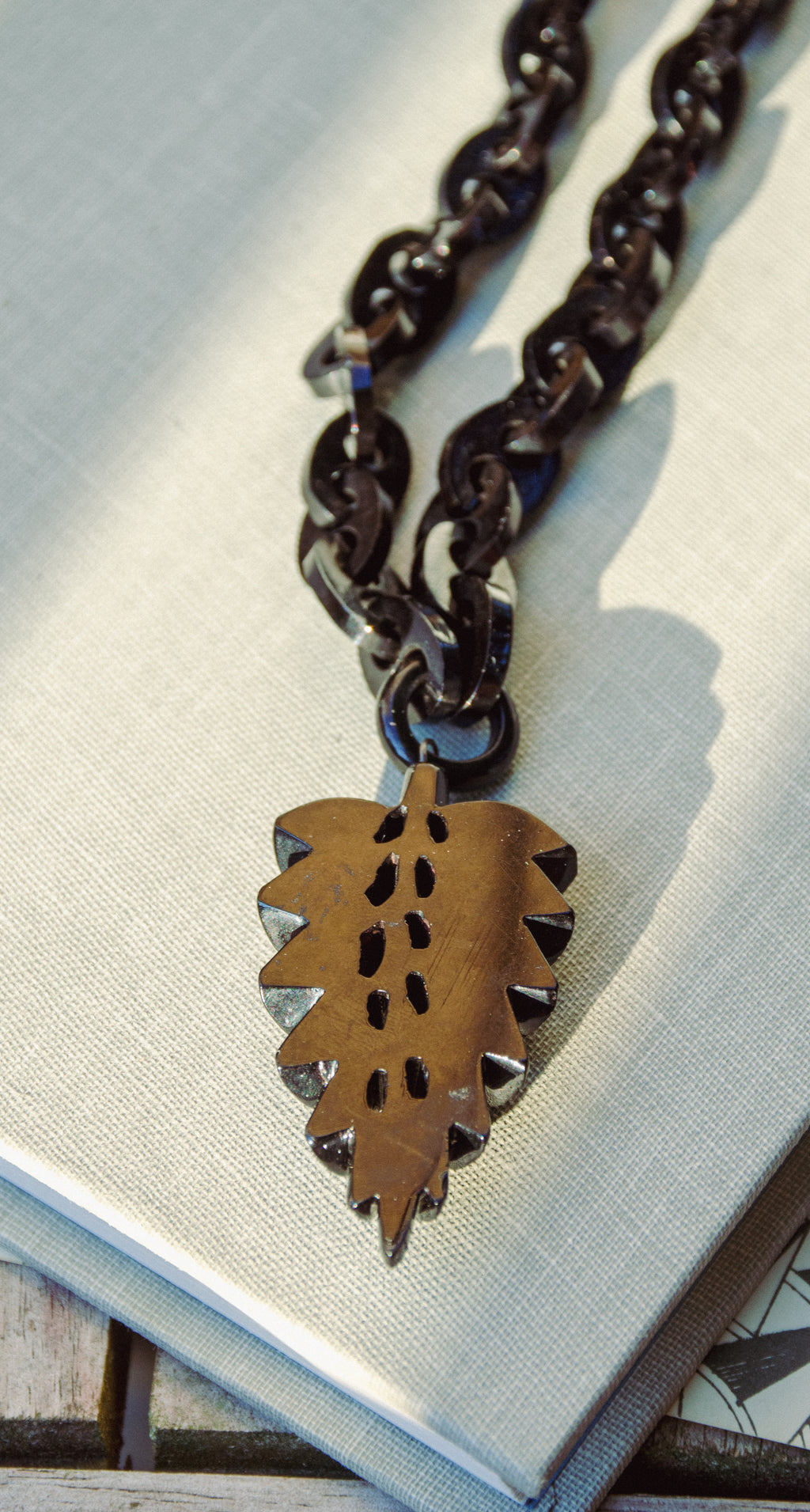 A close-up of a dark metal chain necklace with a leaf-shaped pendant featuring cutout details and small silver-toned accents, resting on a light textured surface.