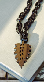 A close-up of a dark metal chain necklace with a leaf-shaped pendant featuring cutout details and small silver-toned accents, resting on a light textured surface.
