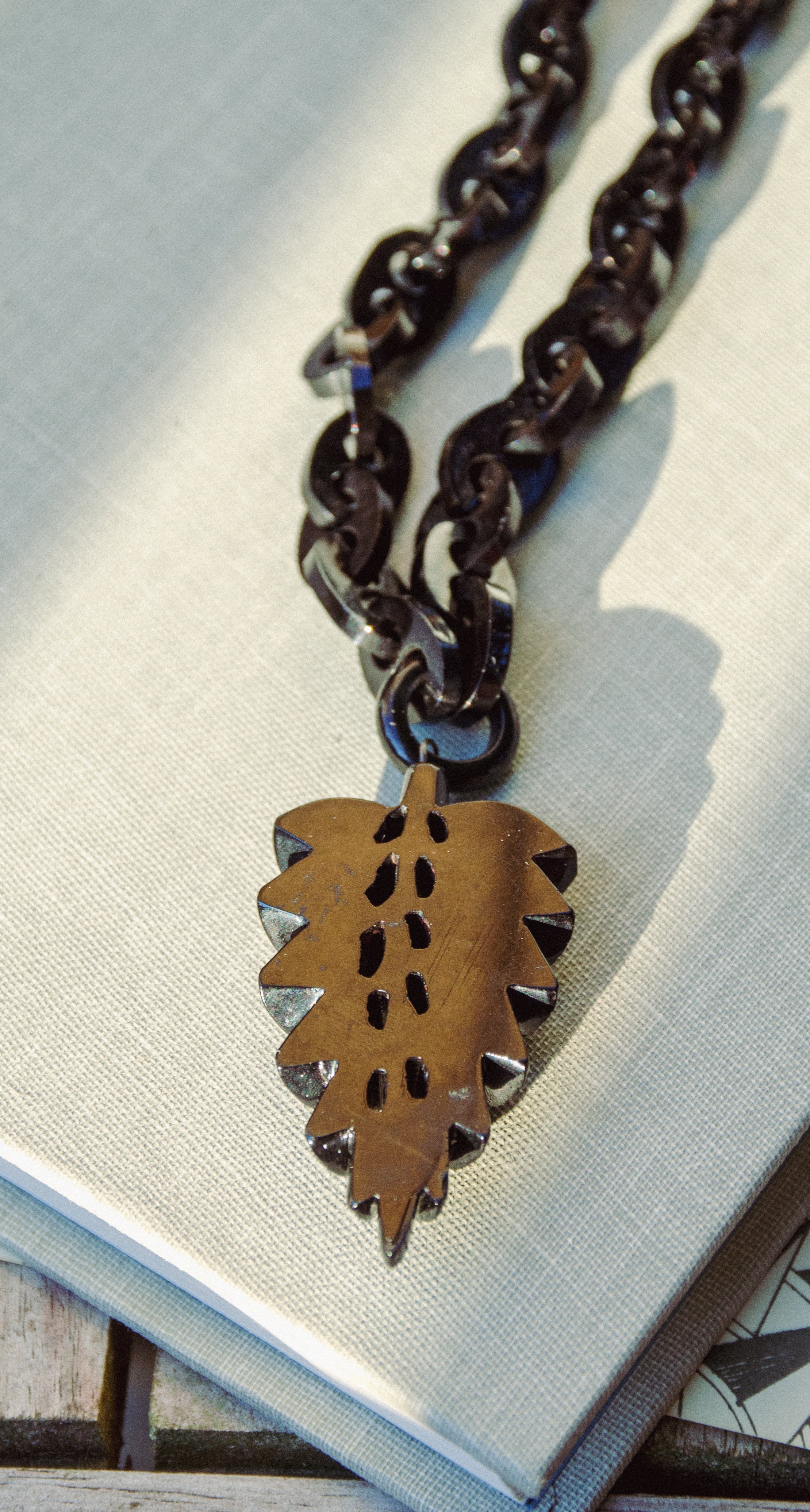 A close-up of a dark metal chain necklace with a leaf-shaped pendant featuring cutout details and small silver-toned accents, resting on a light textured surface.