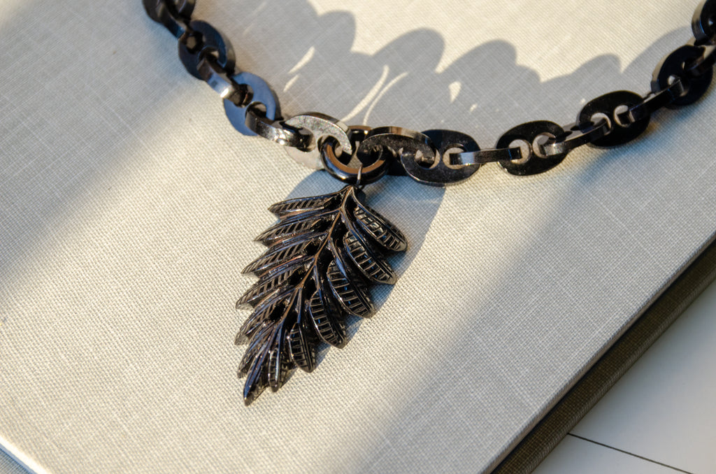 A black Whitby jet mariner chain necklace with a carved fern leaf pendant, displayed on a light gray linen fabric.