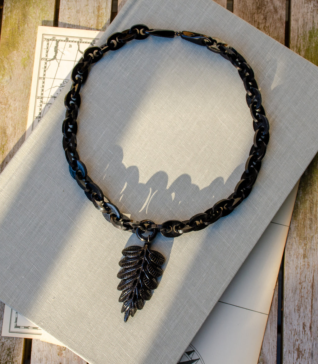 A black Whitby jet marnier chain necklace with a carved fern leaf pendant, displayed on a light gray linen fabric over a rustic wooden surface.