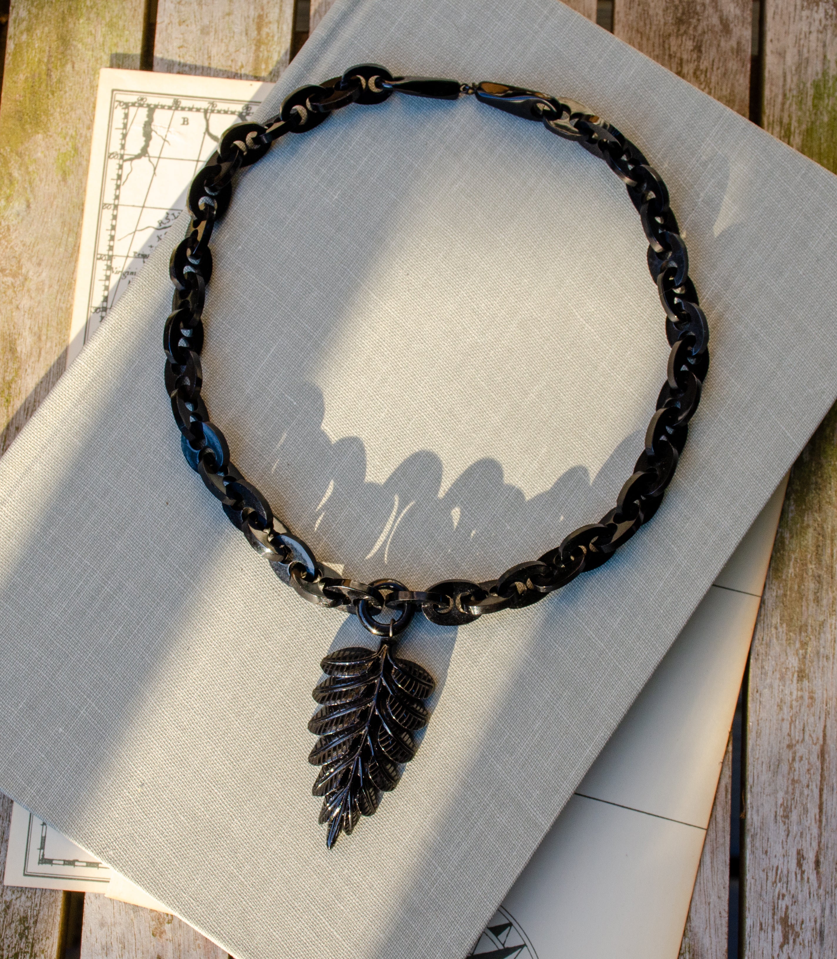 A black Whitby jet marnier chain necklace with a carved fern leaf pendant, displayed on a light gray linen fabric over a rustic wooden surface.