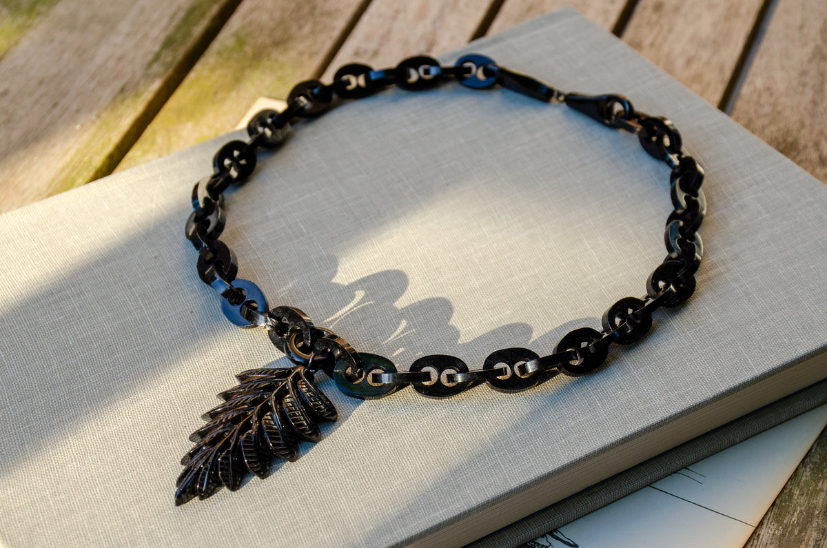 A black Whitby jet marnier chain necklace with a carved fern leaf pendant, displayed on a light gray linen fabric over a rustic wooden surface.