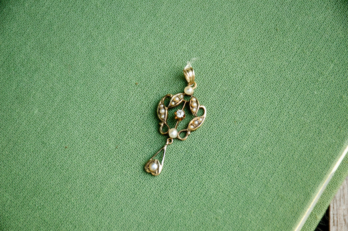 A gold lavalier pendant with seed pearls and a center diamond resting on a green textured surface.