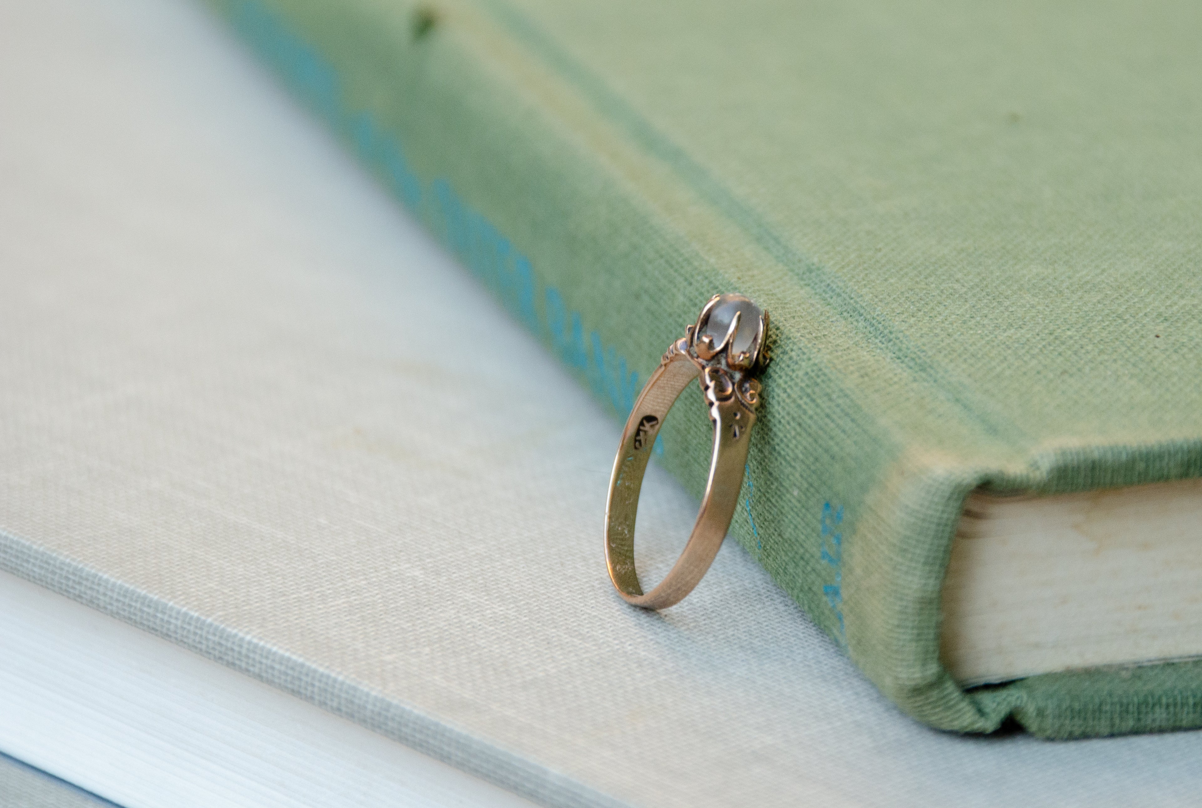 A rose gold ring with an ornate filigree design and a round light blue moonstone, resting on the corner of a light green vintage book.