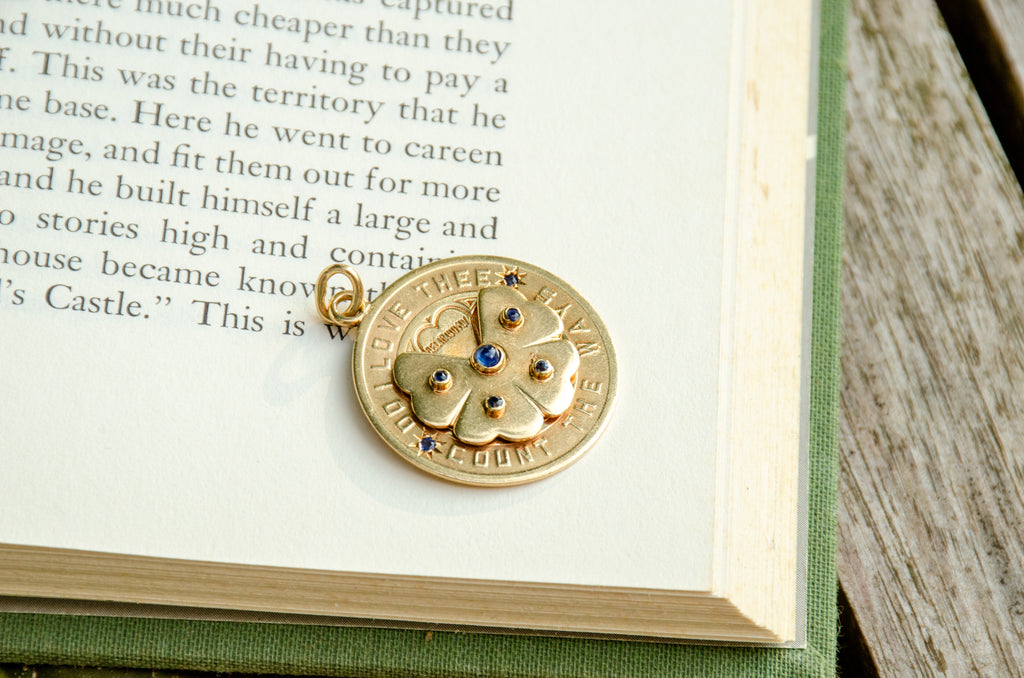 A round gold pendant charm with a floral design and small blue gemstones, resting on an open book page.