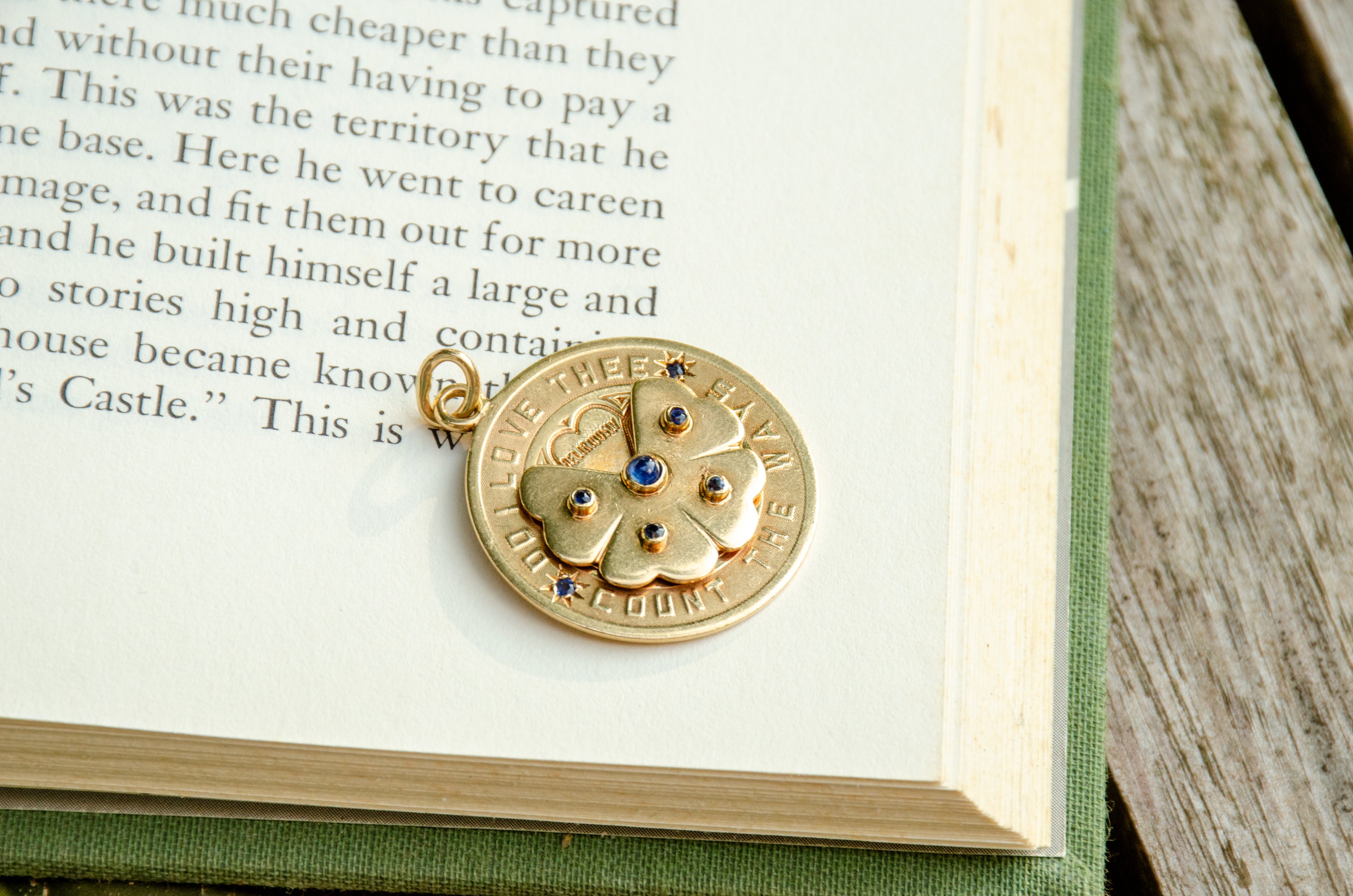 A round gold pendant charm with a floral design and small blue gemstones, resting on an open book page.