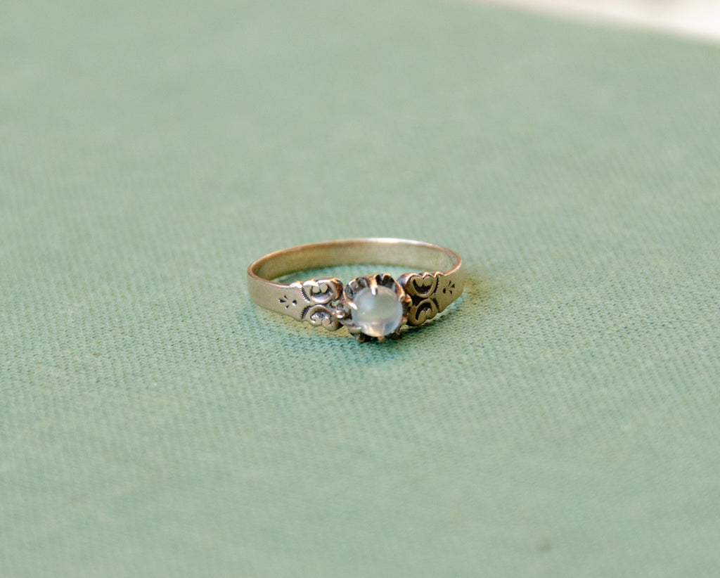 A rose gold ring with an ornate filigree design and a round light blue moonstone, resting on a textured sage green surface.