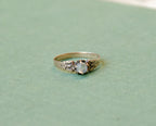A rose gold ring with an ornate filigree design and a round light blue moonstone, resting on a textured sage green surface.