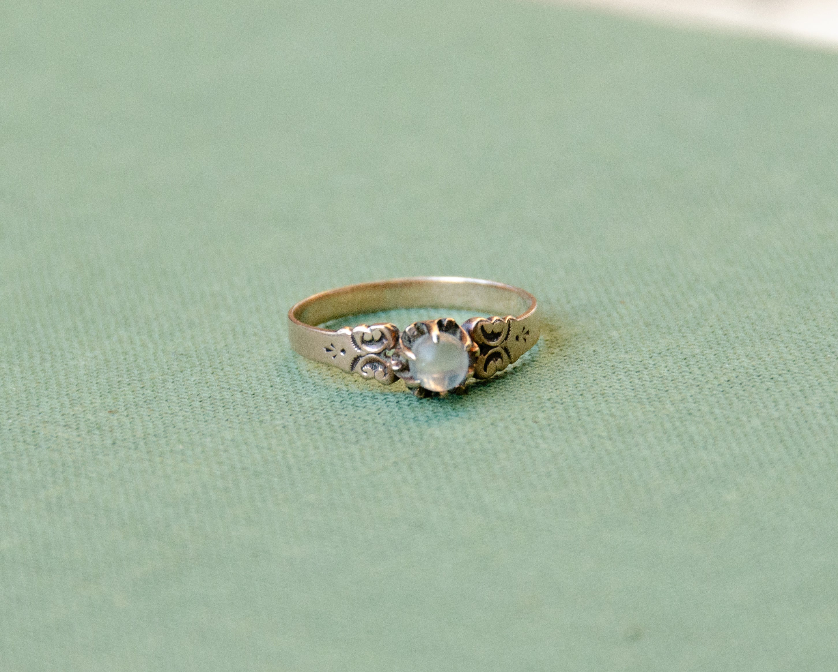 A rose gold ring with an ornate filigree design and a round light blue moonstone, resting on a textured sage green surface.