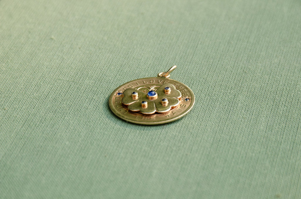 A gold pendant featuring a central flower motif with small blue gemstones, resting on a textured sage green fabric background.