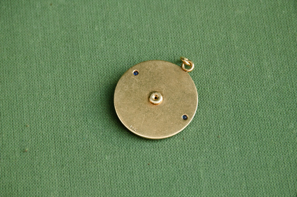 A round hold pendant with a "14k" hallmark resting on a textured green surface.