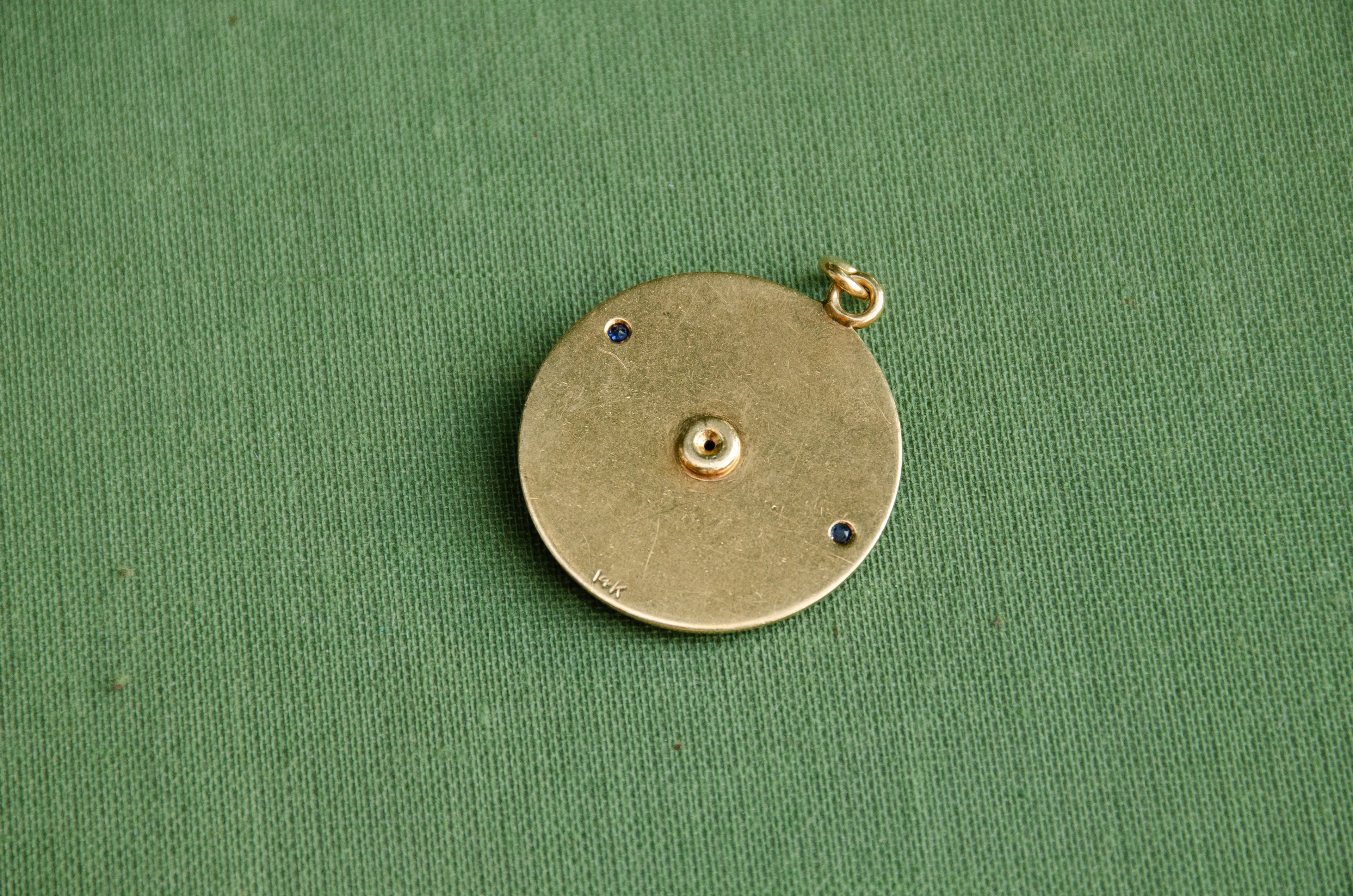 A round hold pendant with a "14k" hallmark resting on a textured green surface.